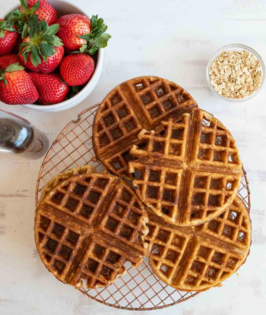 Four golden-brown waffles on a cooling rack. A bowl of fresh strawberries, a small bowl of oats, and a bottle of syrup are next to them, set on a light wooden surface.