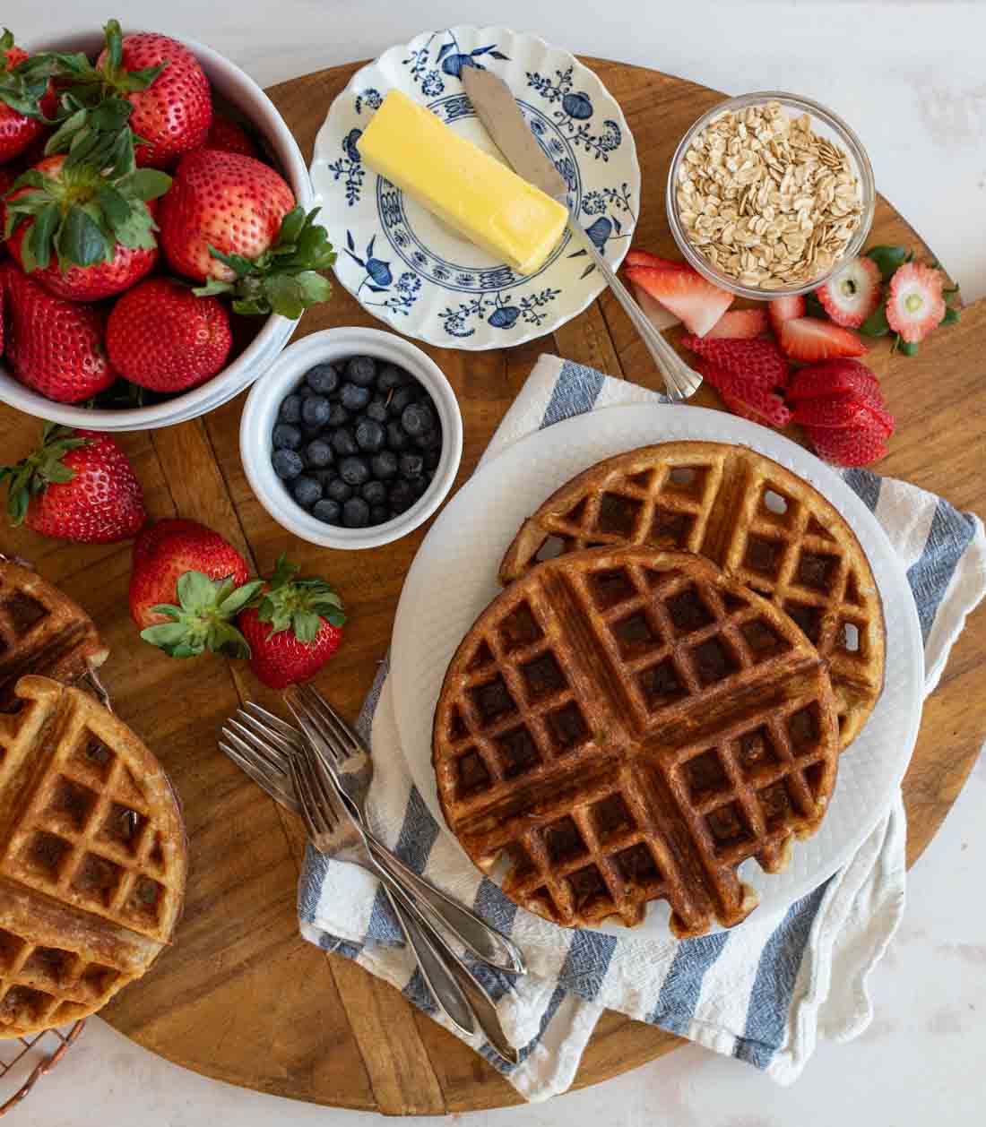 A wooden board with fresh strawberries, blueberries, oats, butter, and waffles on a plate. Forks rest beside the plate, and a striped napkin enhances the presentation.