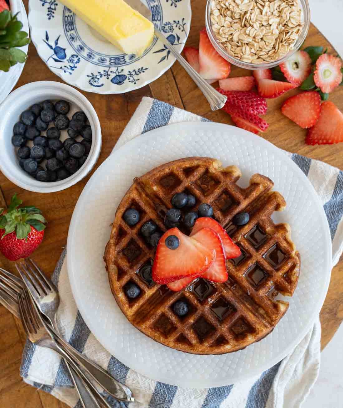 Oatmeal waffles topped with sliced strawberries and blueberries sits on a white plate. Nearby, theres a dish of blueberries, a butter dish with butter, a bowl of oats, sliced strawberries, and silverware on a striped napkin, all on a wooden table.