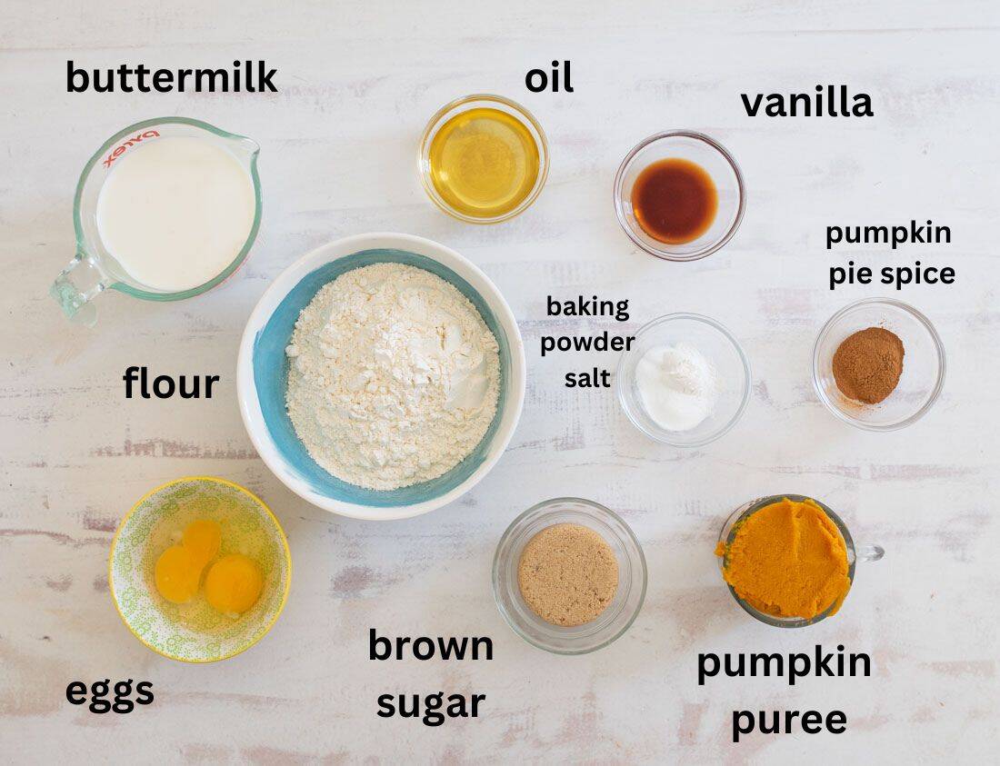 A flat lay of ingredients for baking includes buttermilk, oil, vanilla, flour, baking powder, salt, pumpkin pie spice, eggs, brown sugar, and pumpkin puree. Each ingredient is labeled and displayed in small bowls or a measuring cup.