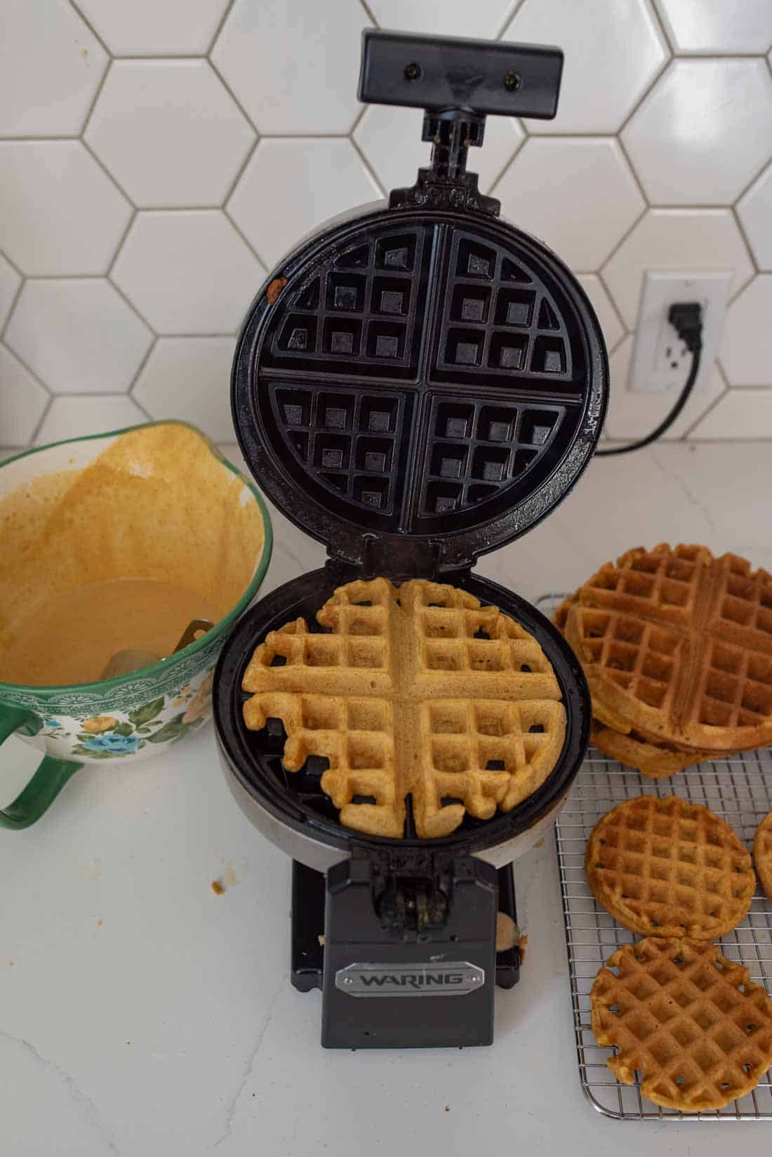 A waffle maker is open, showing freshly made waffles inside. Several waffles are cooling on a wire rack nearby. A mixing bowl with waffle batter is on the counter next to the waffle maker, against a hexagonal tile backsplash.