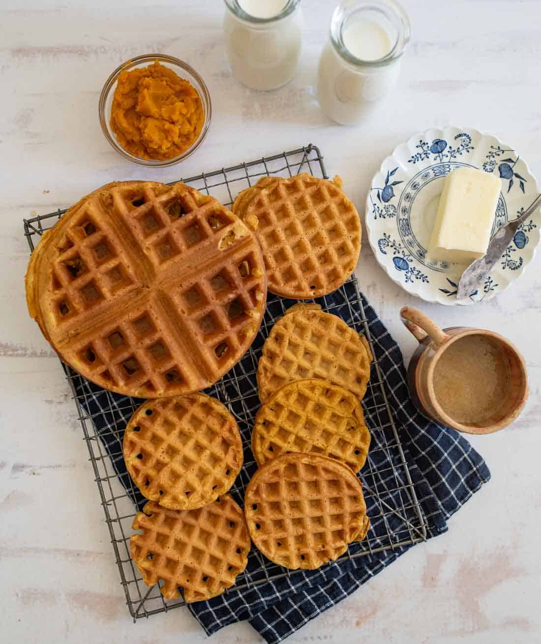 Top view of various waffles on a cooling rack, alongside a jar of pumpkin puree, a plate with butter and a knife, two bottles of milk, and a mug of coffee. The setup is on a white surface with a blue cloth beneath the rack.