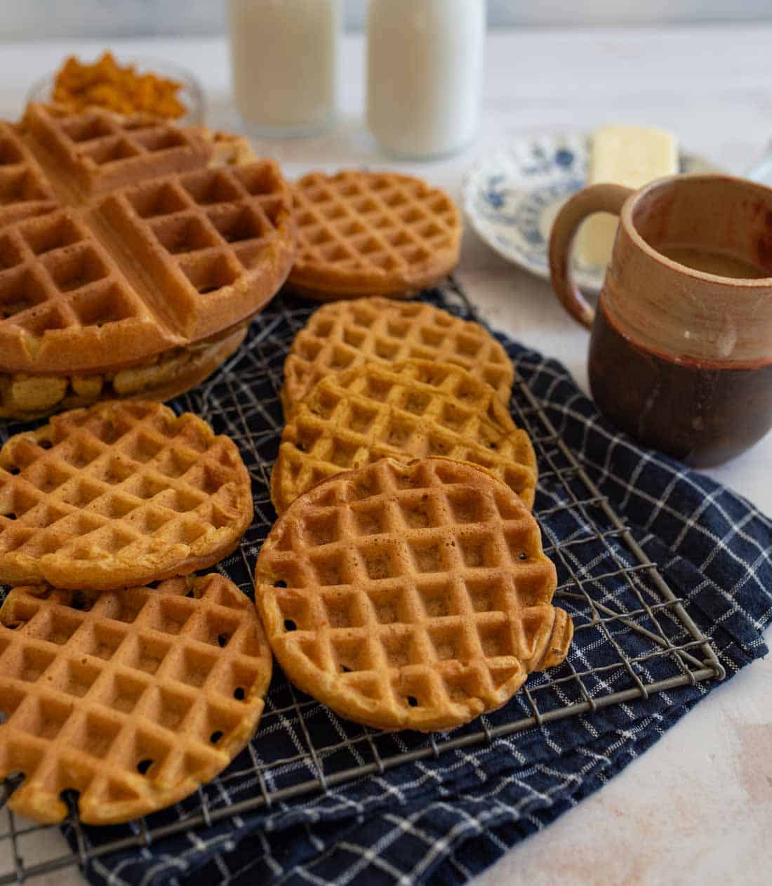 A variety of waffles are displayed on a dark checkered cloth over a cooling rack. A mug, a plate with butter, and two bottles in the background complete the breakfast setting.