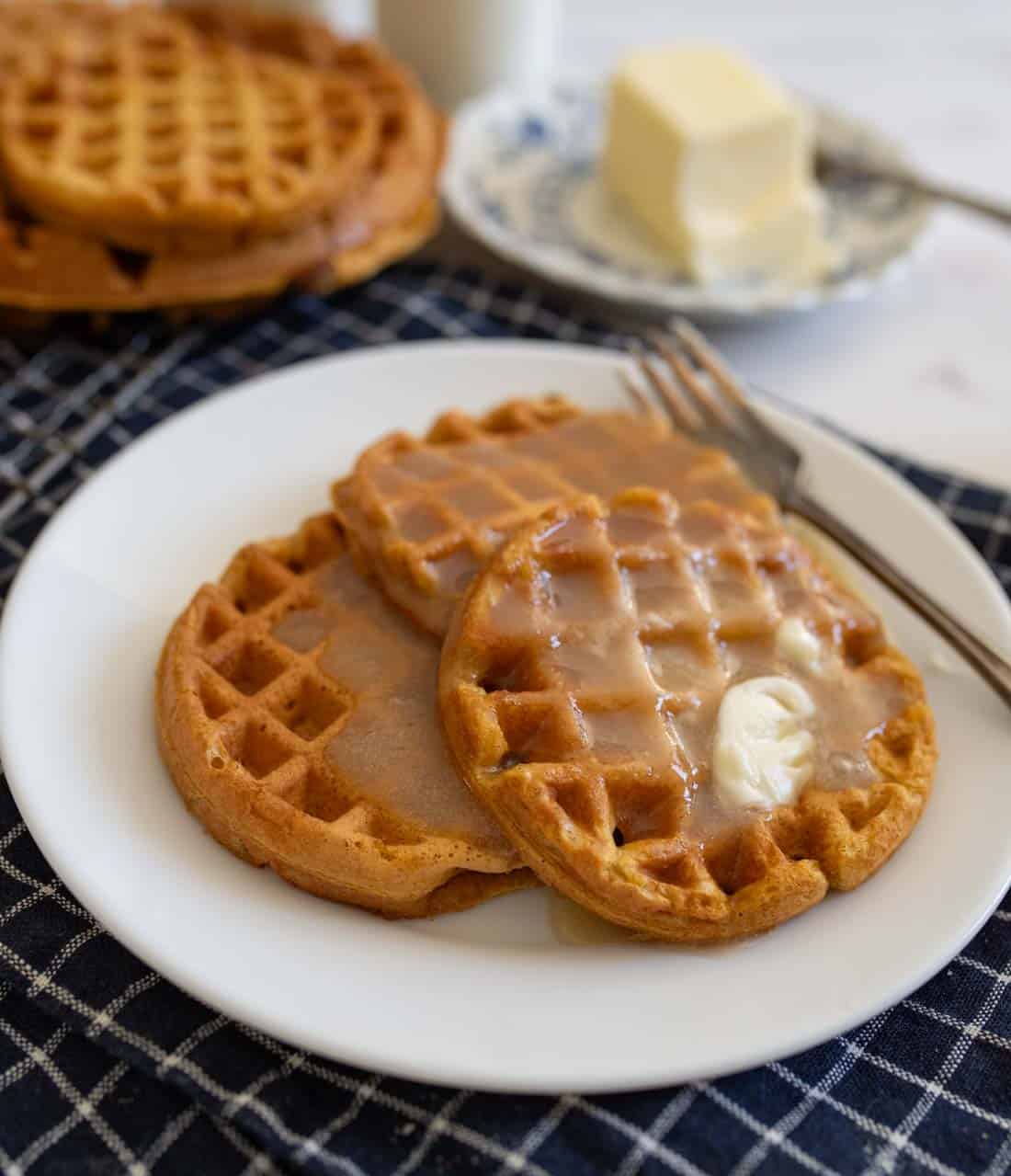 Two pumpkin waffles on a white plate, drizzled with syrup and topped with butter. A fork rests nearby. In the background, a stack of waffles and a dish with a butter block sit on a checkered cloth.