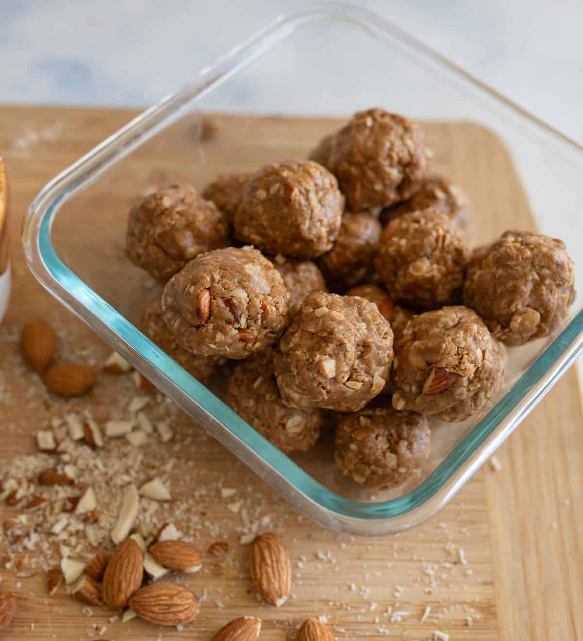 A glass container filled with almond butter protein balls made of oats, nuts, and other ingredients sits on a wooden surface. Whole and chopped almonds are scattered beside the container.