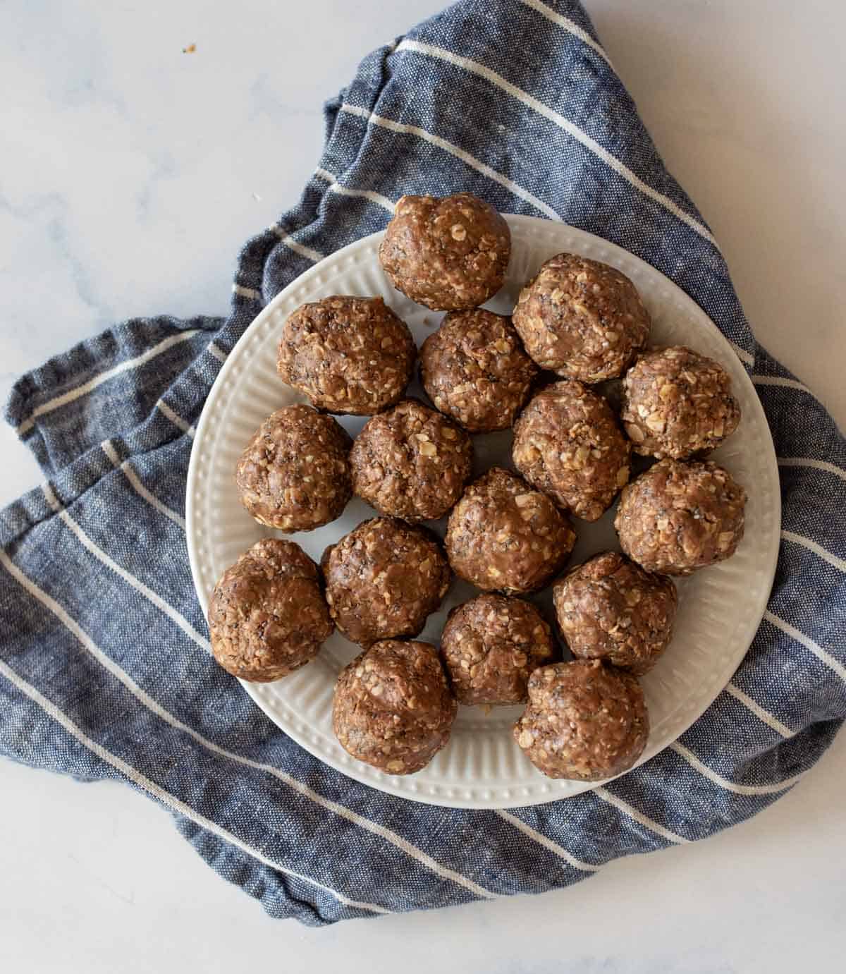 A white plate with several round, brown chocolate peanut butter protein balls topped with oats. The plate is placed on a crumpled, blue and white striped cloth on a marble surface.
