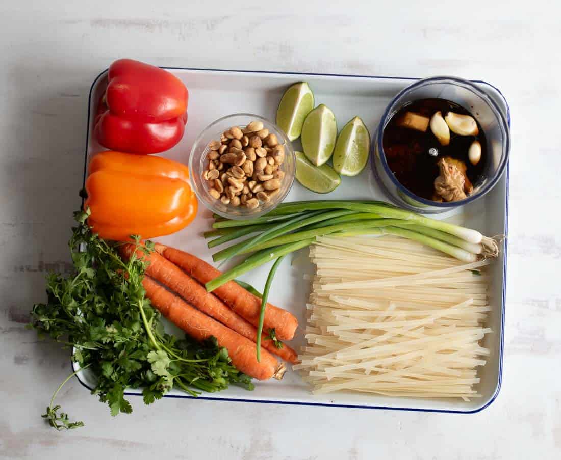 A tray with rice noodles, green onions, carrots, cilantro, red and orange bell peppers, lime wedges, peanuts, and a bowl of sauce with garlic and tofu, all arranged neatly on a white surface.