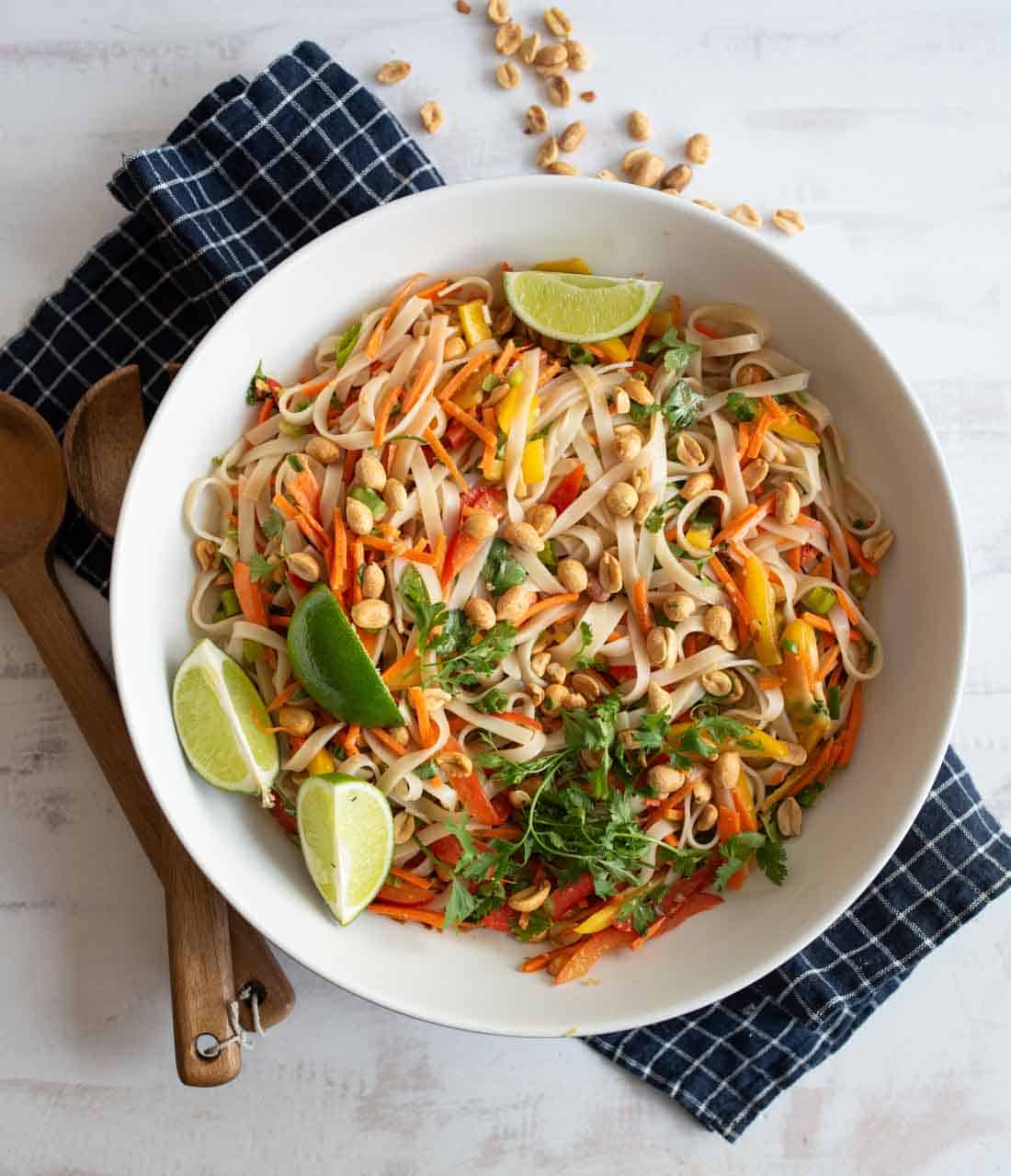 A white bowl of colorful Asian noodle salad with carrots, peppers, peanuts, cilantro, and lime wedges sits on a blue checkered napkin, with wooden utensils nearby. Some peanuts are scattered on the table.