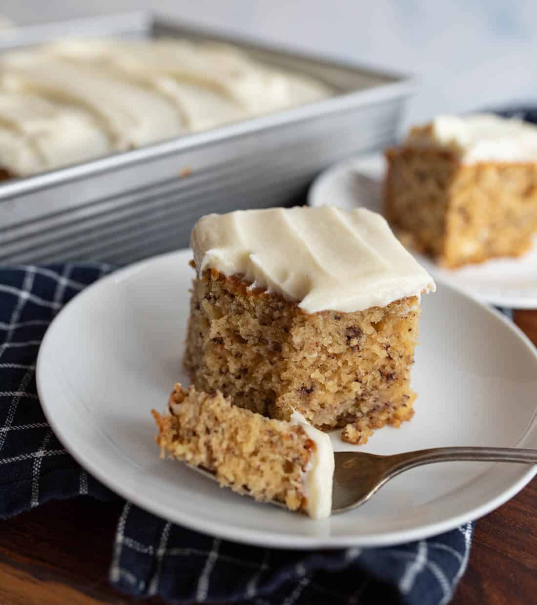 A square slice of banana cake with cream cheese frosting sits on a white plate with a fork holding a bite, in front of a pan and another plate with more cake. A dark checkered cloth is underneath the plate.