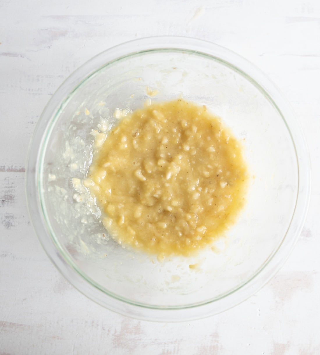 A clear glass bowl containing mashed bananas sits on a white surface, with the banana mixture appearing soft and slightly lumpy.