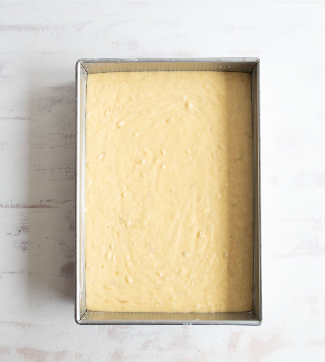 A rectangular metal baking pan filled with smooth, pale yellow cake batter sits on a white surface.