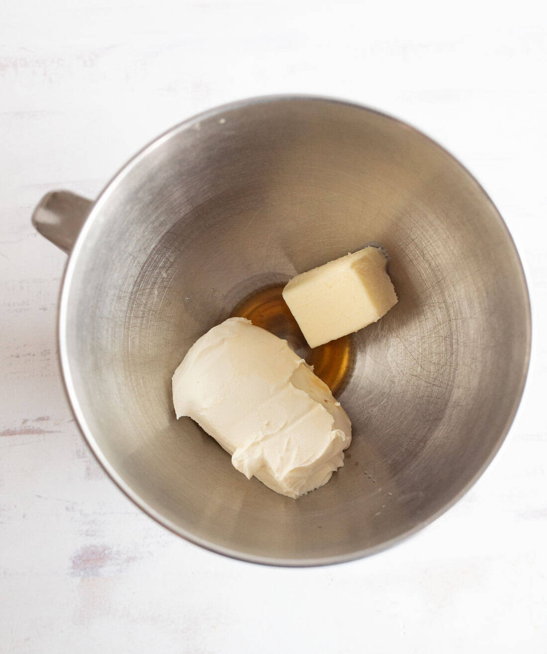 A metal mixing bowl containing a block of cream cheese, a small piece of butter, and a small amount of vanilla extract.