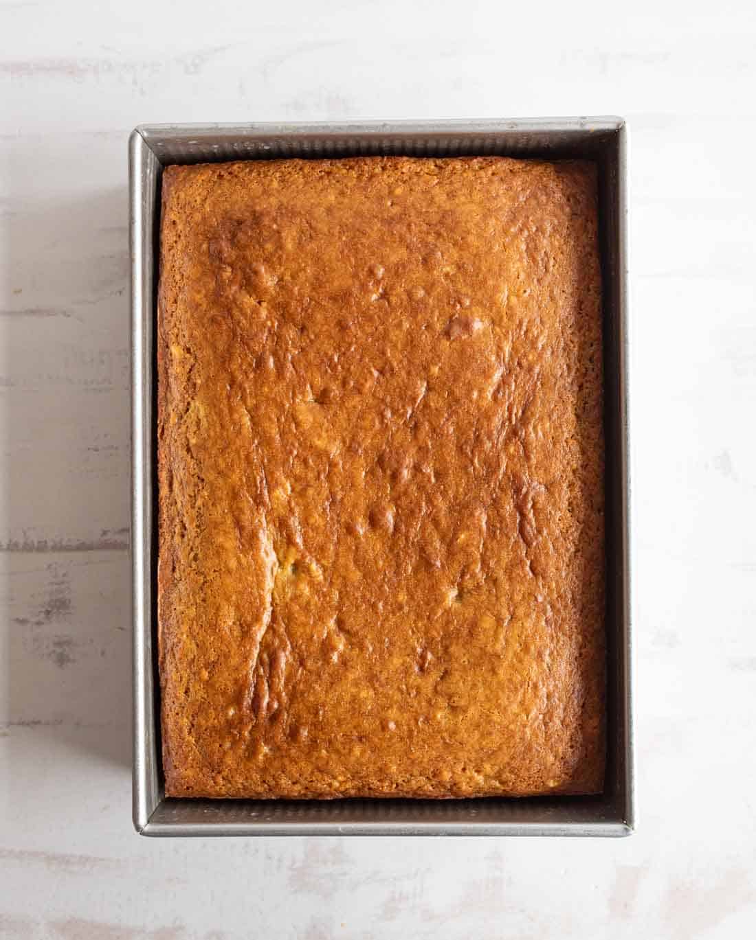 A golden-brown, rectangular loaf of baked banana bread sits in a metal baking pan on a light-colored surface.