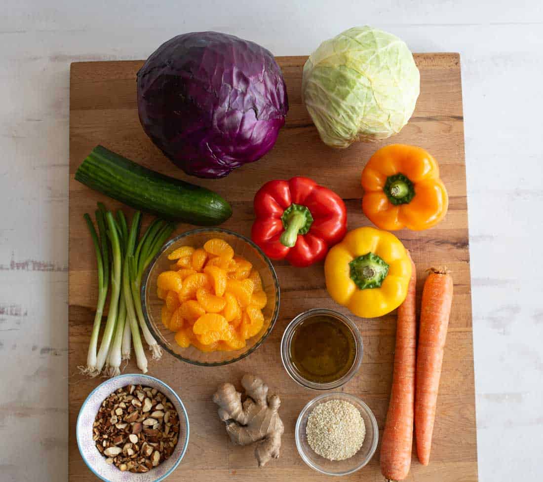 A wooden board displays red and green cabbage, cucumber, green onions, a bowl of mandarin oranges, red and yellow bell peppers, carrots, fresh ginger, sesame seeds, a bowl of dressing, and a bowl of mixed nuts.