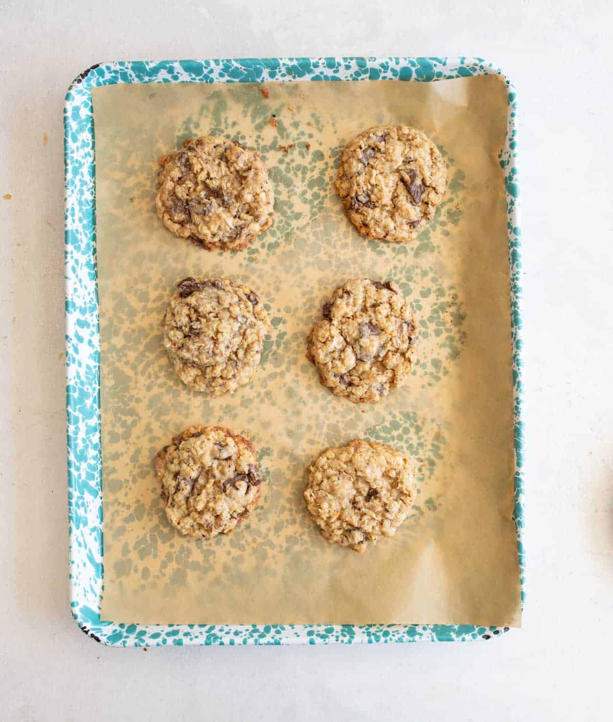 A blue speckled baking tray lined with parchment paper holds six oatmeal chocolate chip cookies arranged in two columns of three on a light-colored surface.
