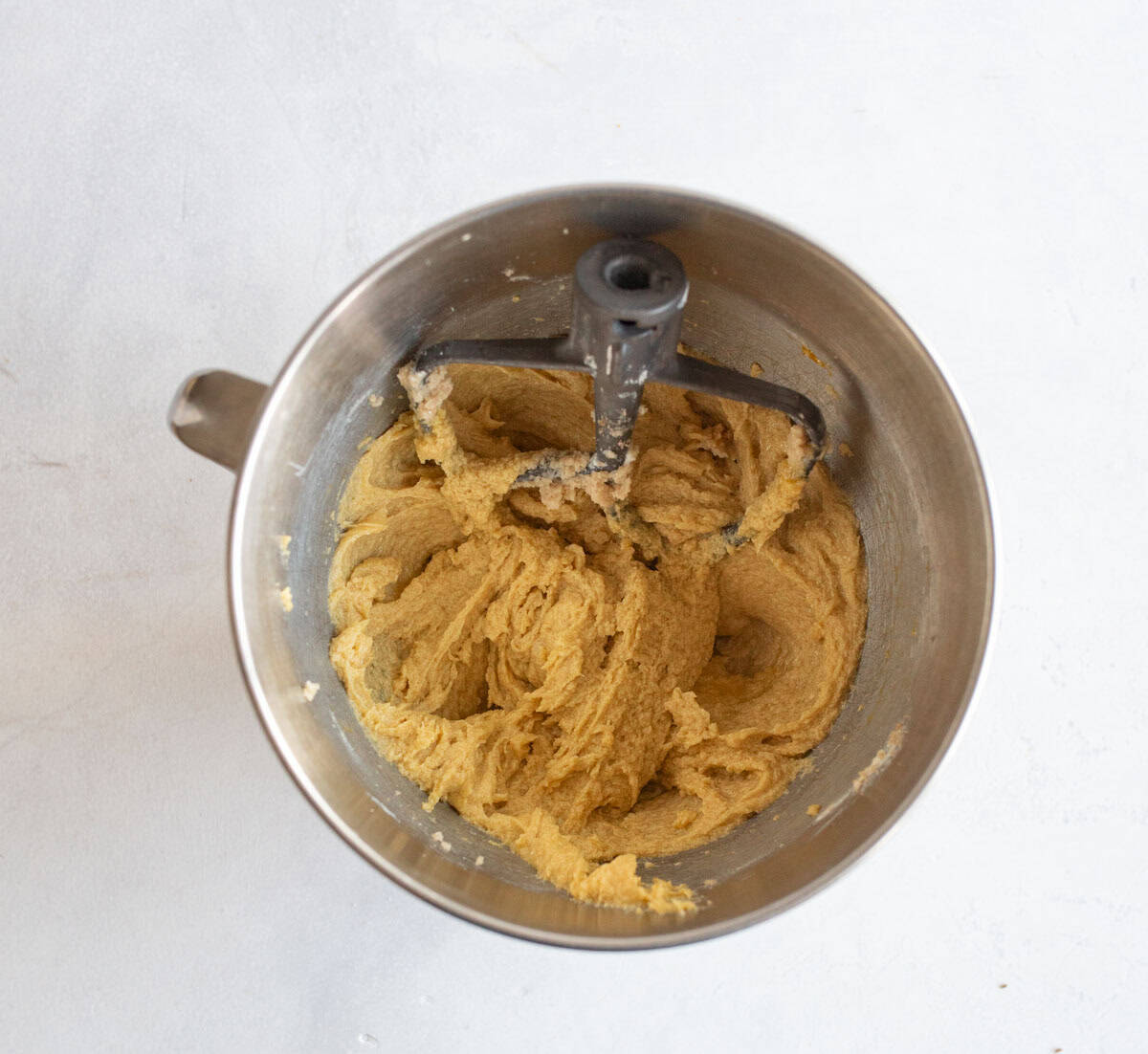 A mixing bowl with a paddle attachment, filled with light brown cookie dough, sits on a white surface.