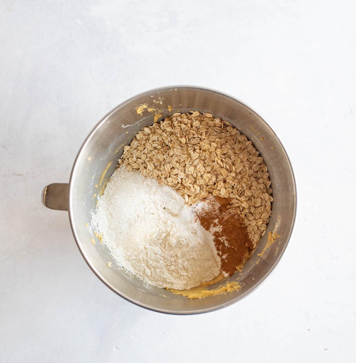 A mixing bowl containing oats, flour, cinnamon, and sugar on a light surface, viewed from above.