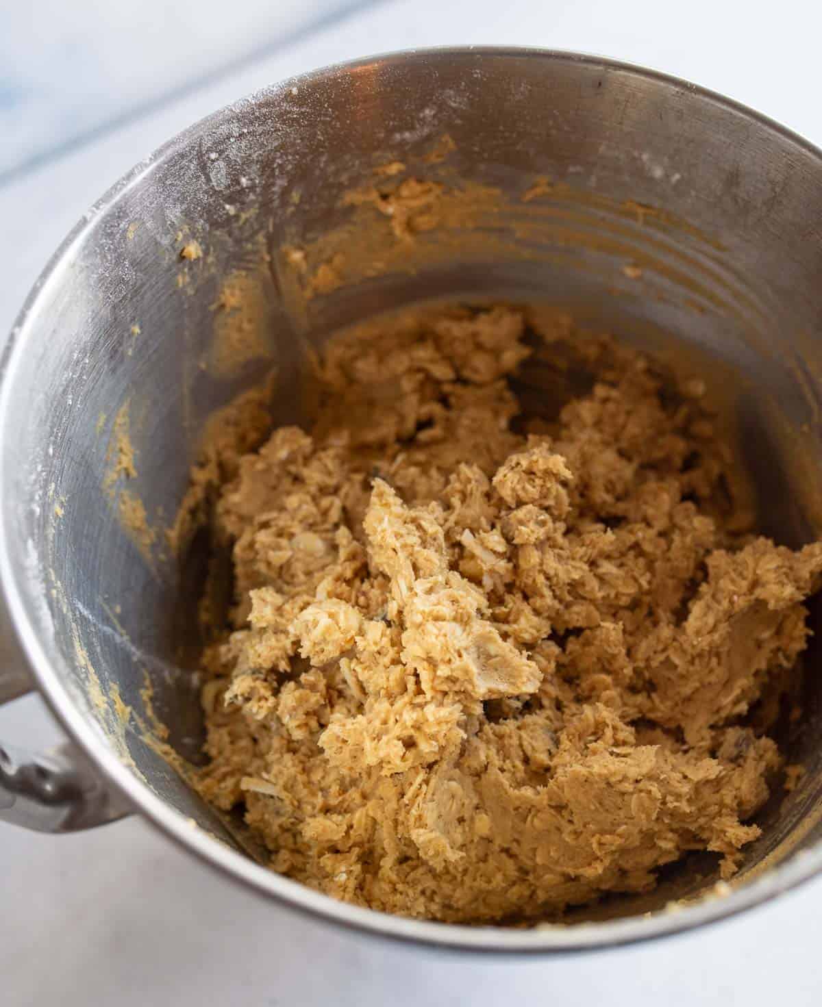 A stainless steel mixing bowl containing thick, light brown cookie dough with visible oats, sitting on a white countertop.
