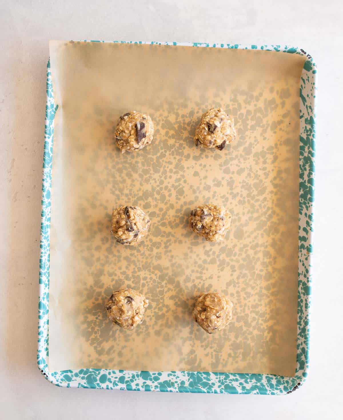A teal-speckled baking sheet lined with parchment paper holds six evenly spaced balls of cookie dough, ready to be baked.