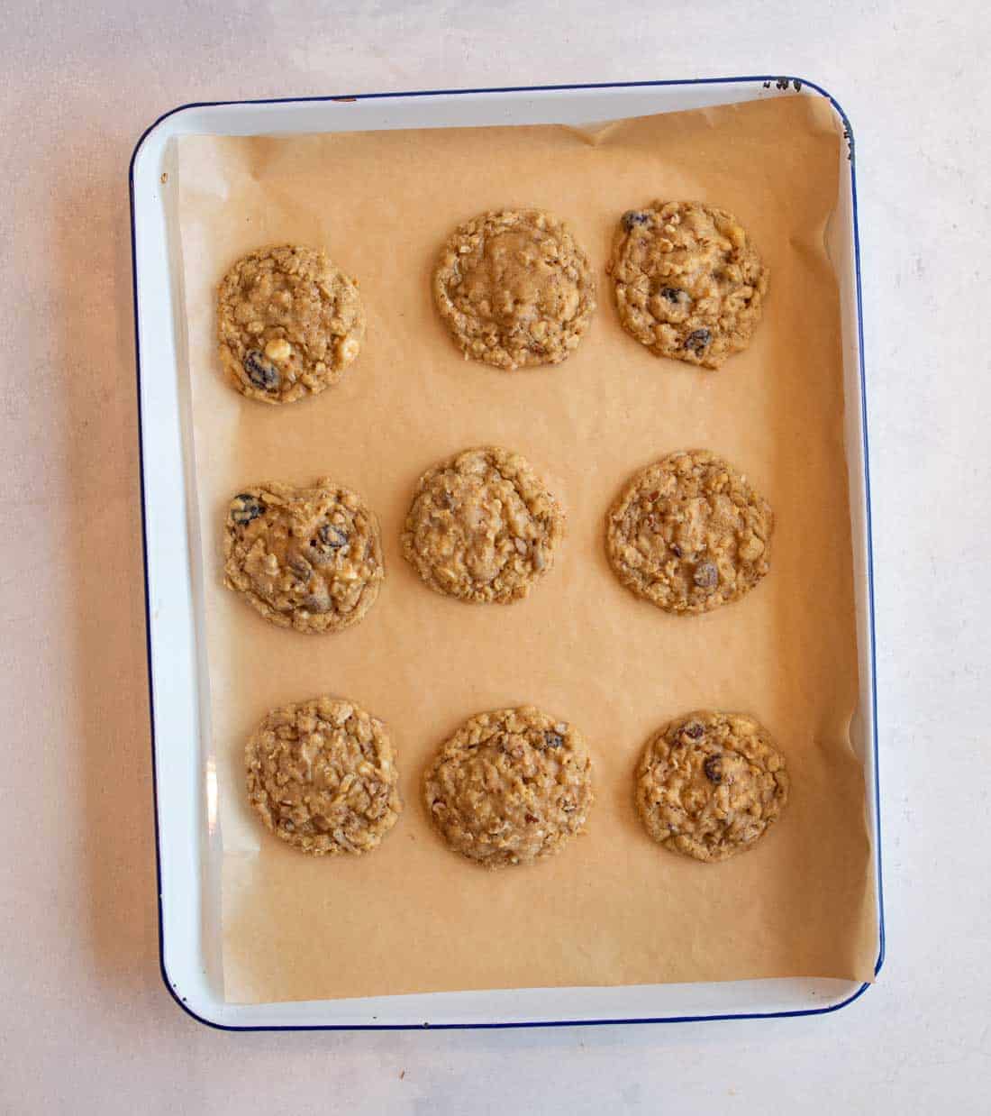 A white baking tray lined with parchment paper holds nine unbaked oatmeal cookies spaced evenly apart, ready to be baked.