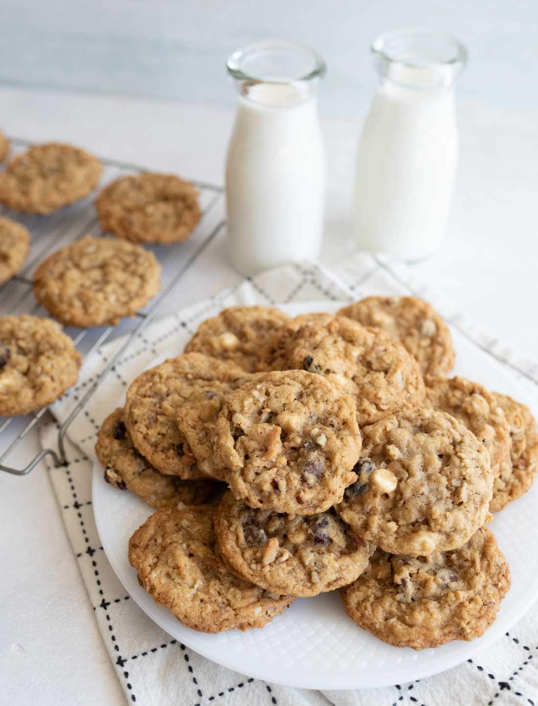 A plate of cowboy oatmeal cookies with chocolate chips and white chocolate chips sits on a checkered cloth next to two bottles of milk; more cookies cool on a wire rack in the background.
