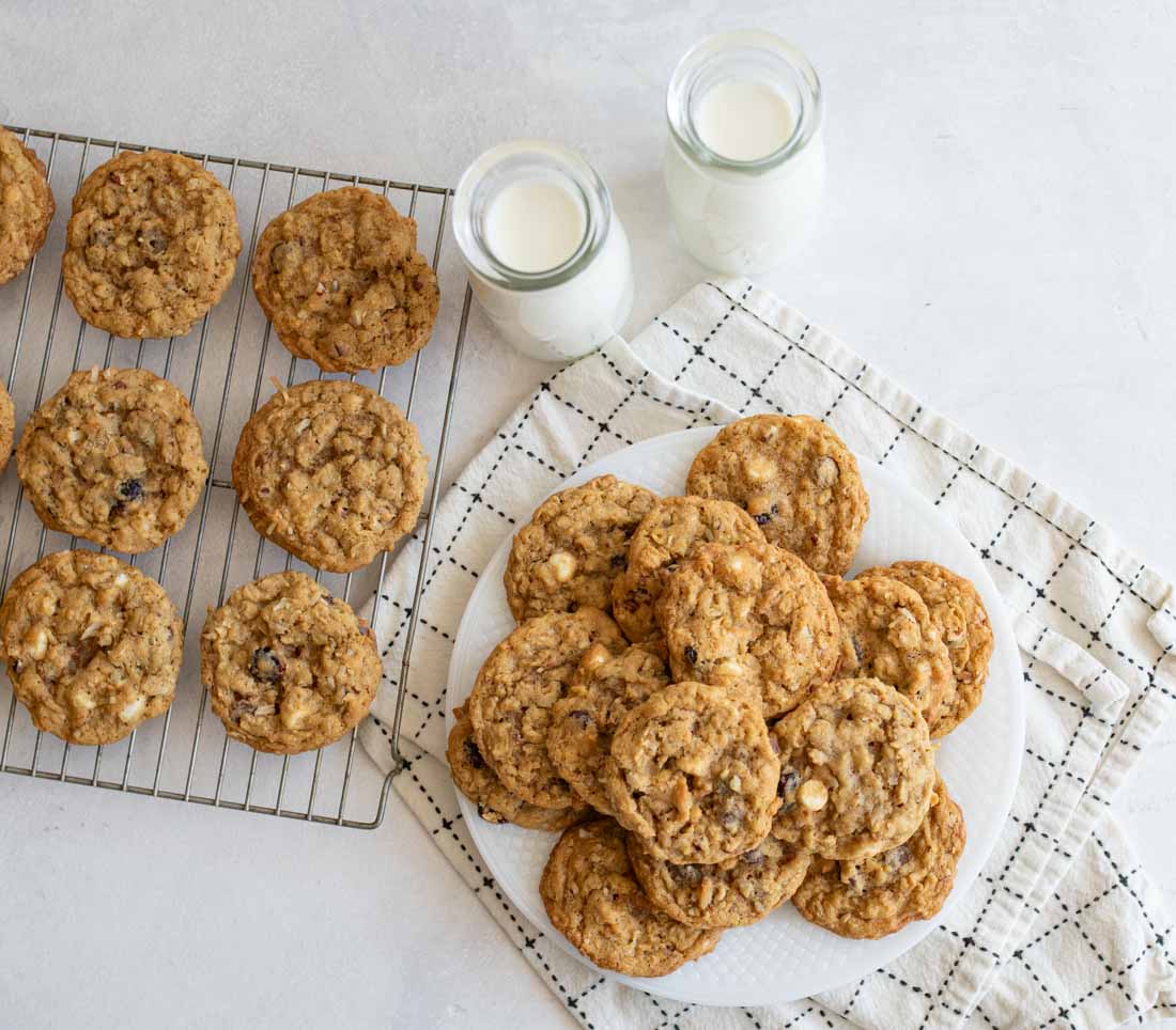 A plate of oatmeal cookies sits on a white towel next to two glasses of milk. More cookies are cooling on a wire rack to the side. The background is a light-colored surface.