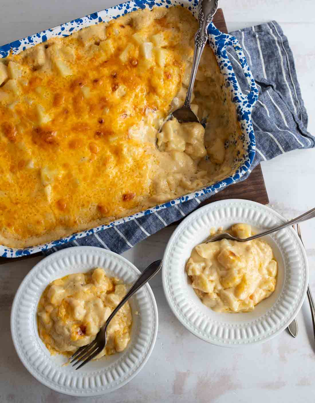 A blue and white speckled baking dish filled with cheesy potatoes, partially served into two white bowls with forks, all set on a striped cloth.