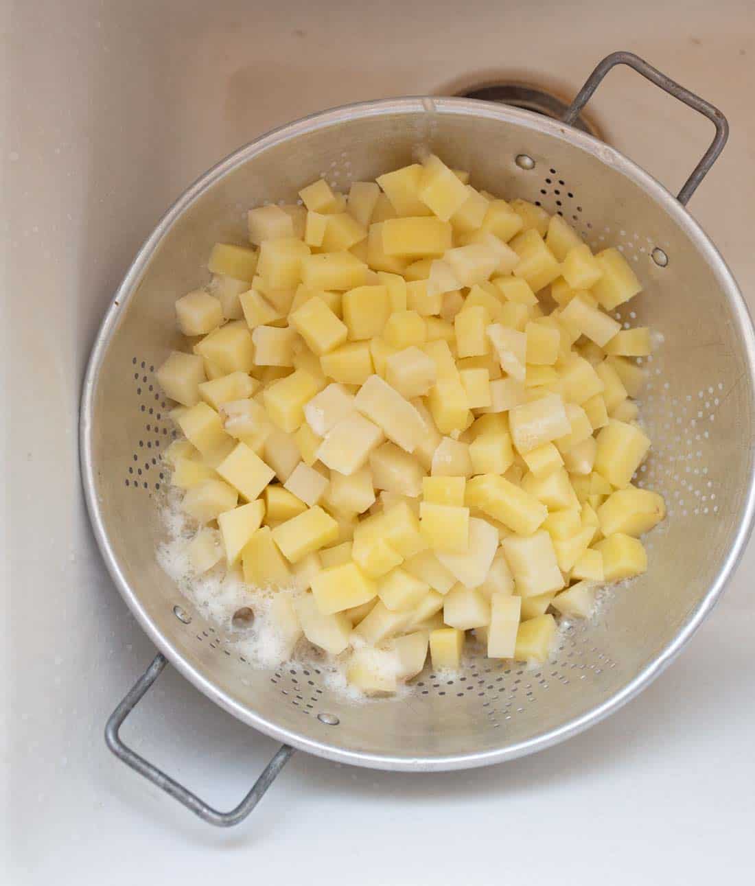 A metal colander filled with small, evenly diced raw potatoes sits in a white sink, with some water and foam visible around the potatoes.