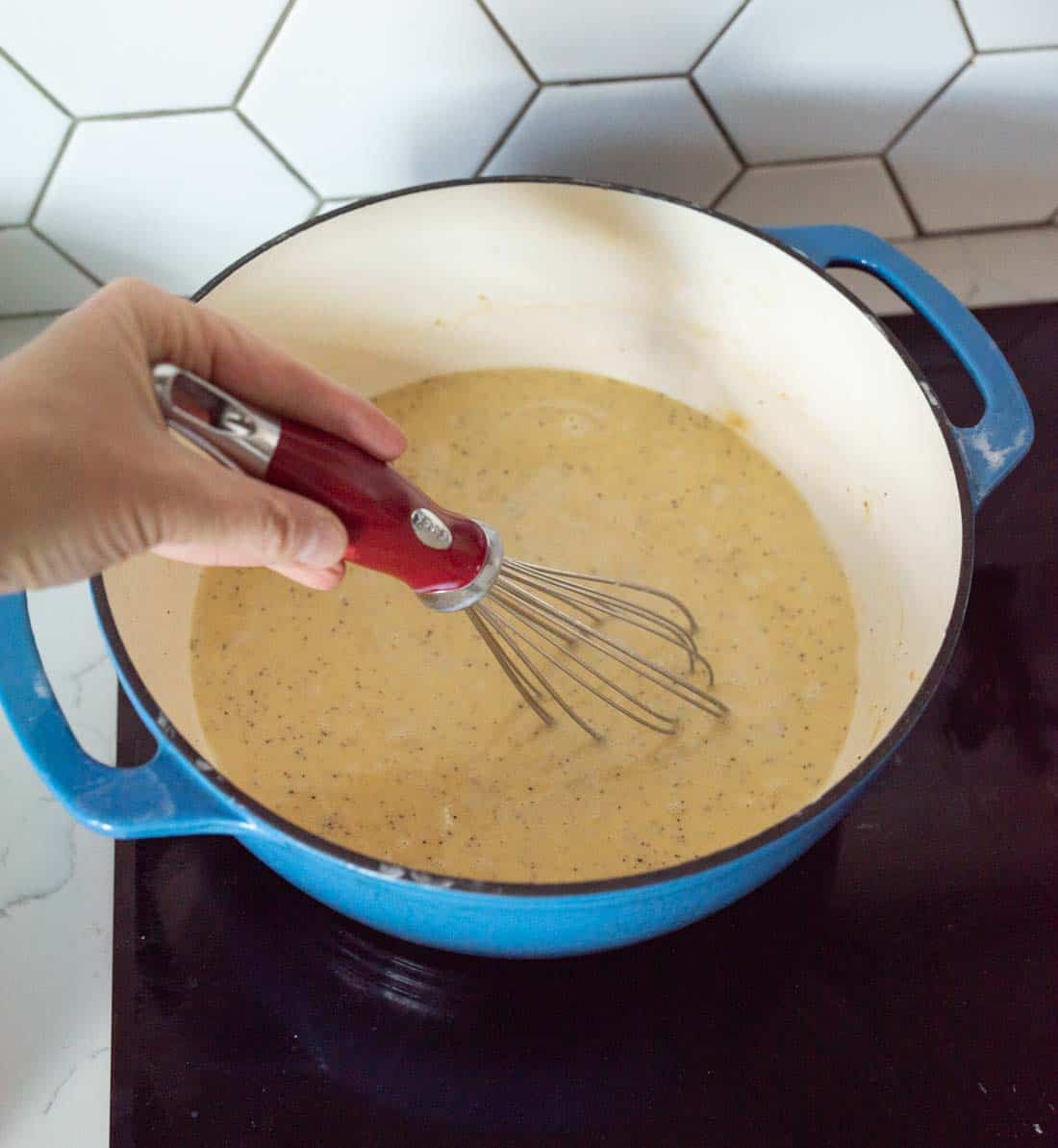 A hand uses a whisk to stir a creamy sauce in a blue pot on a stovetop, with a white hexagon-tiled backsplash visible in the background.