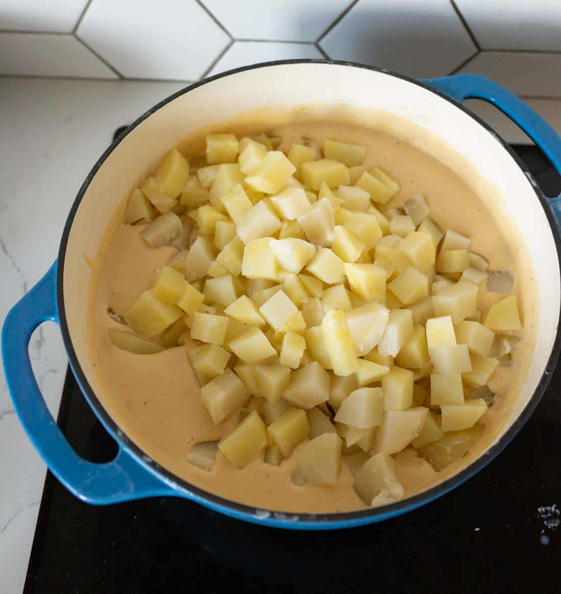 A blue pot filled with creamy sauce and topped with a layer of diced potatoes sits on a black stovetop in a kitchen with white hexagon tile backsplash.