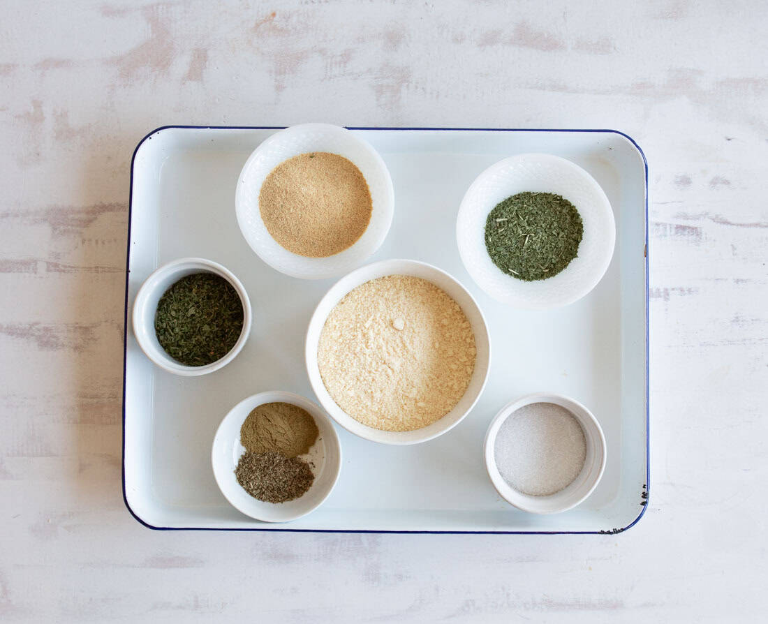 A white tray holds six small white bowls filled with various dried herbs and powders, arranged neatly on a light-colored surface.