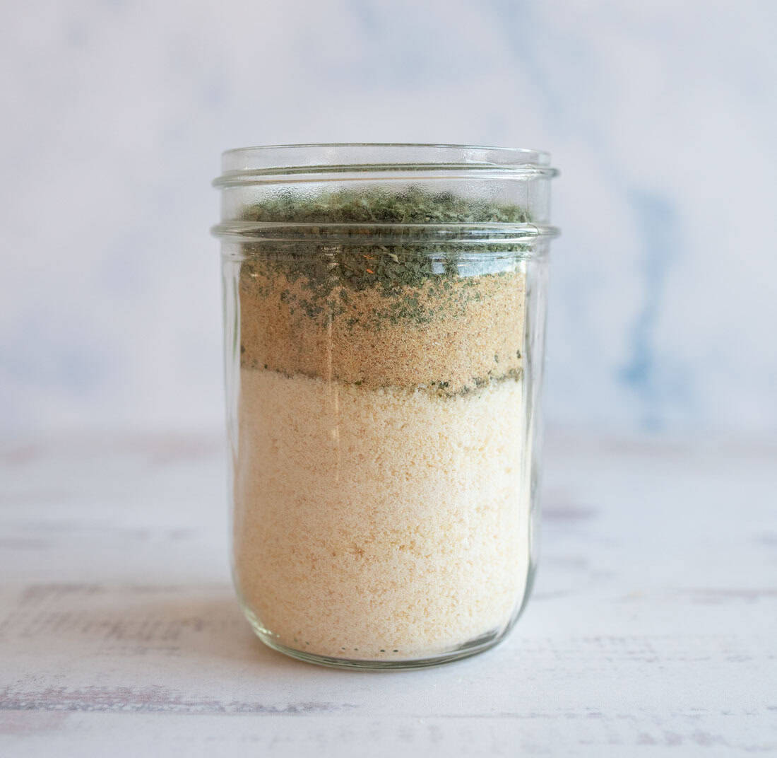 A glass mason jar filled with layers of dry ingredients, including a white powdery substance on the bottom, a light brown layer in the middle, and a green herb layer on top, set against a light background.