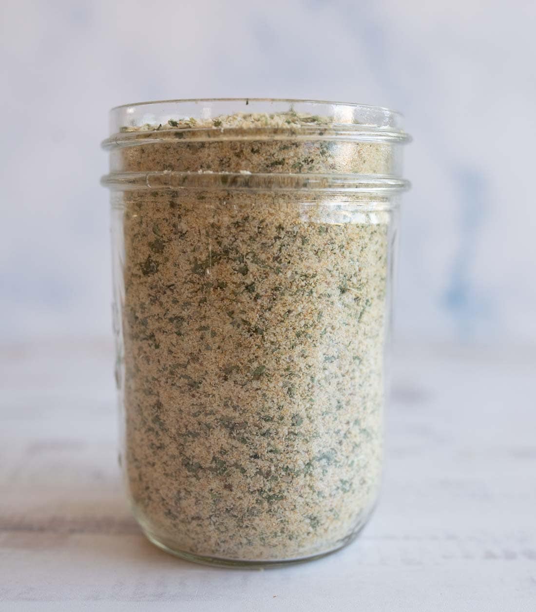 A glass jar filled with garlic parmesan seasoning, showing a blend of fine crumbs and green herbs, placed on a light-colored surface with a soft, blurred background.