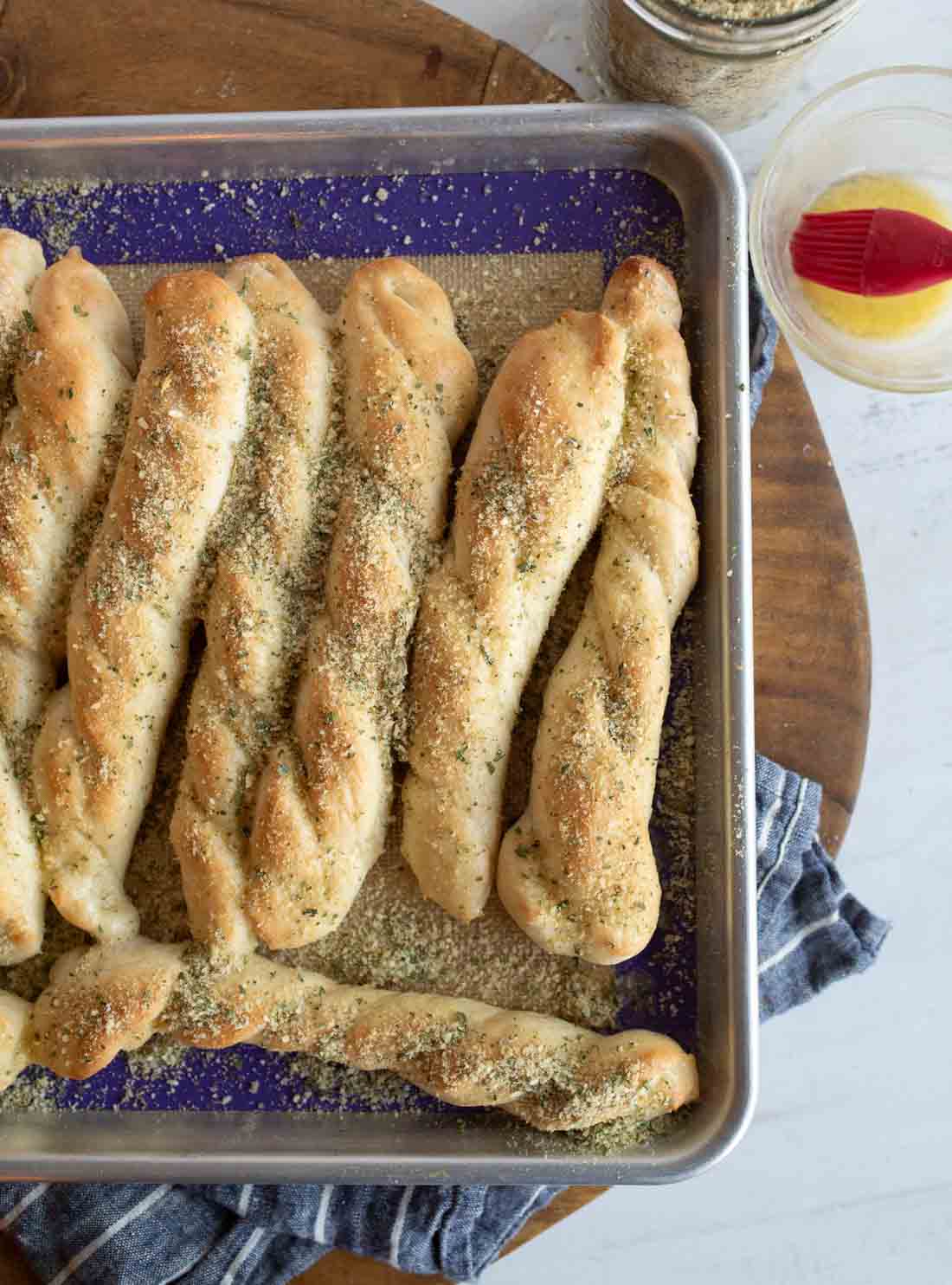 A baking sheet with several golden-brown, twisted breadsticks sprinkled with garlic parmesan seasoning, resting on a blue cloth on a wooden surface. A jar with a red brush and seasoning is nearby.