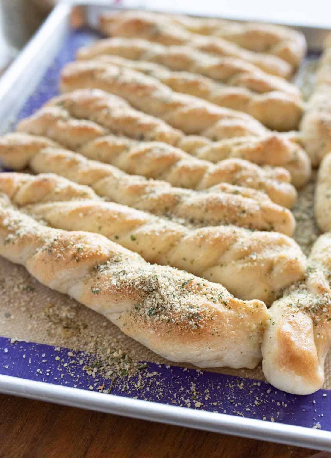 A baking tray filled with freshly baked twisted breadsticks, sprinkled with herbs and grated cheese, resting on a parchment-lined surface.