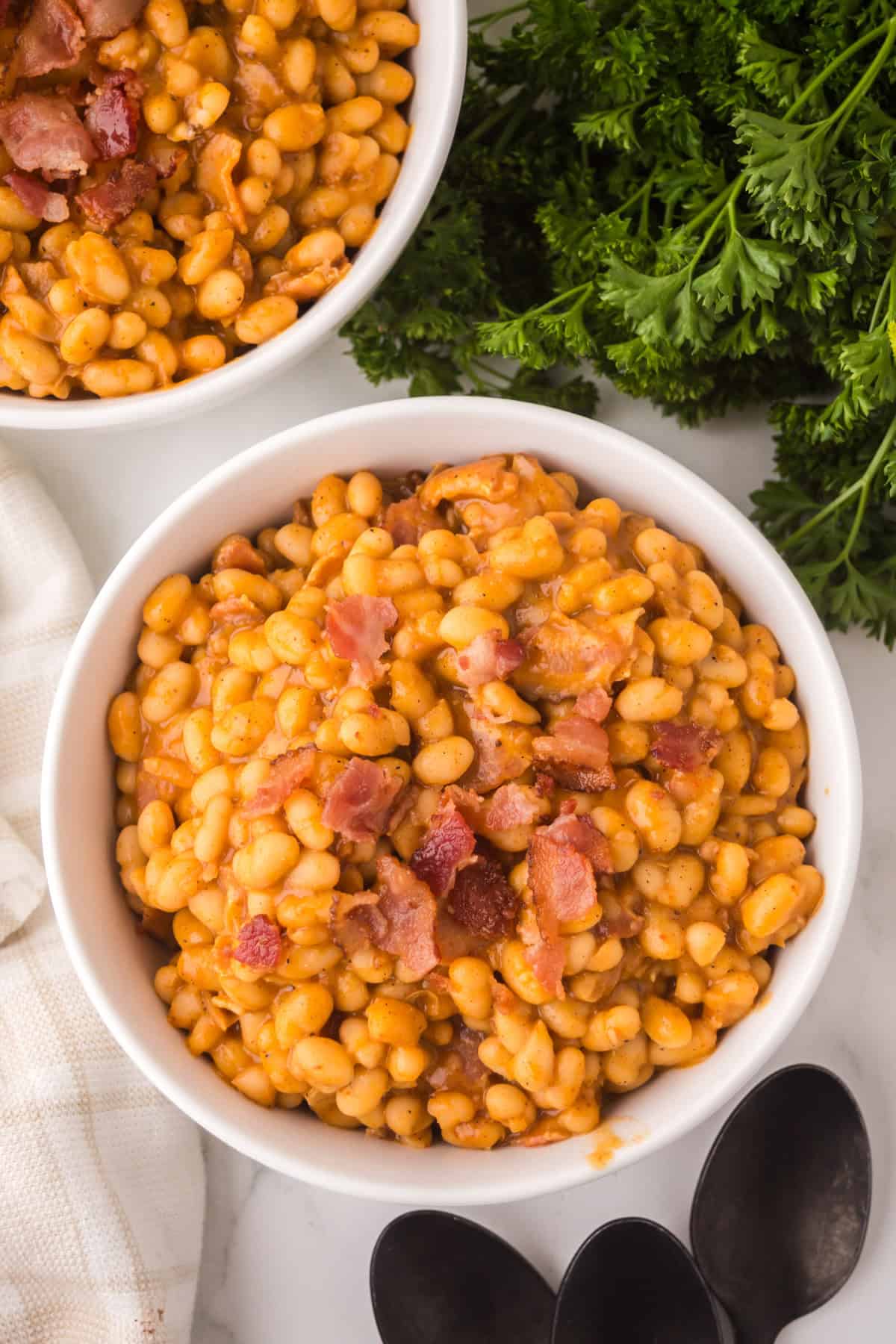 A white bowl filled with homemade baked beans and pieces of bacon, placed on a white surface next to fresh parsley, a white cloth, black spoons, and part of another bowl of beans.