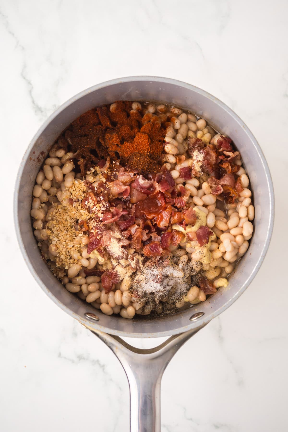 A saucepan on a white marble surface filled with homemade baked beans, crumbled bacon, minced garlic, spices, and seasonings, all arranged in separate sections before being mixed together.