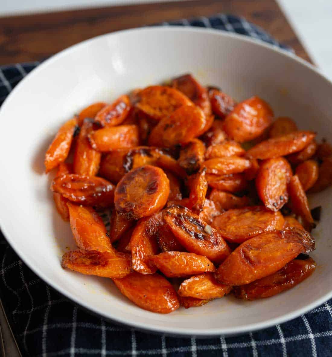 A white bowl filled with honey roasted carrots sits on a dark, checkered cloth. The carrots are glazed and slightly caramelized, giving them a shiny, golden-brown appearance.