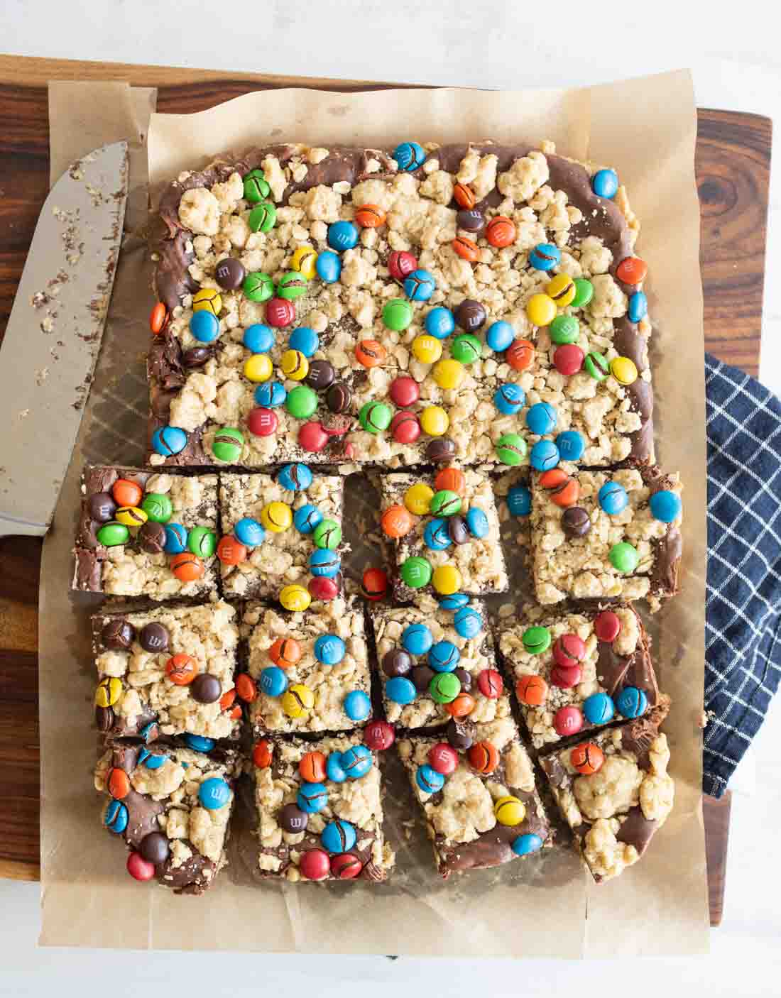 A tray of chocolate oat bars topped with colorful candy-coated chocolates, partially sliced on parchment paper with a large knife and a blue plaid cloth nearby.