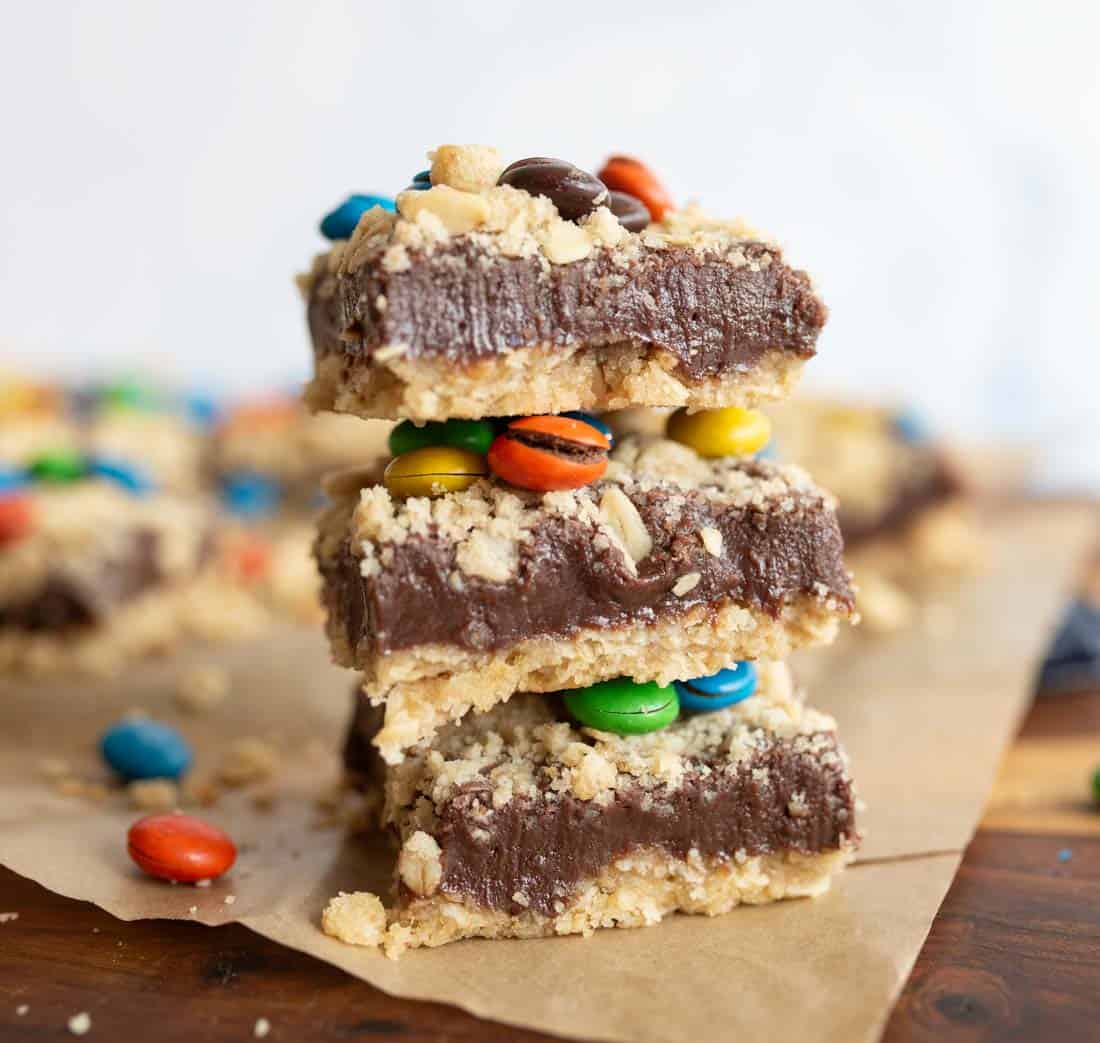 Three chocolate fudge bars with a crumbly topping and colorful candy-coated chocolates are stacked on parchment paper, with more bars and candies blurred in the background.