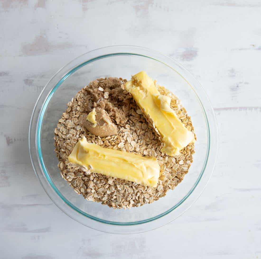 A clear glass bowl containing rolled oats, brown sugar, and two sticks of butter on a white surface.