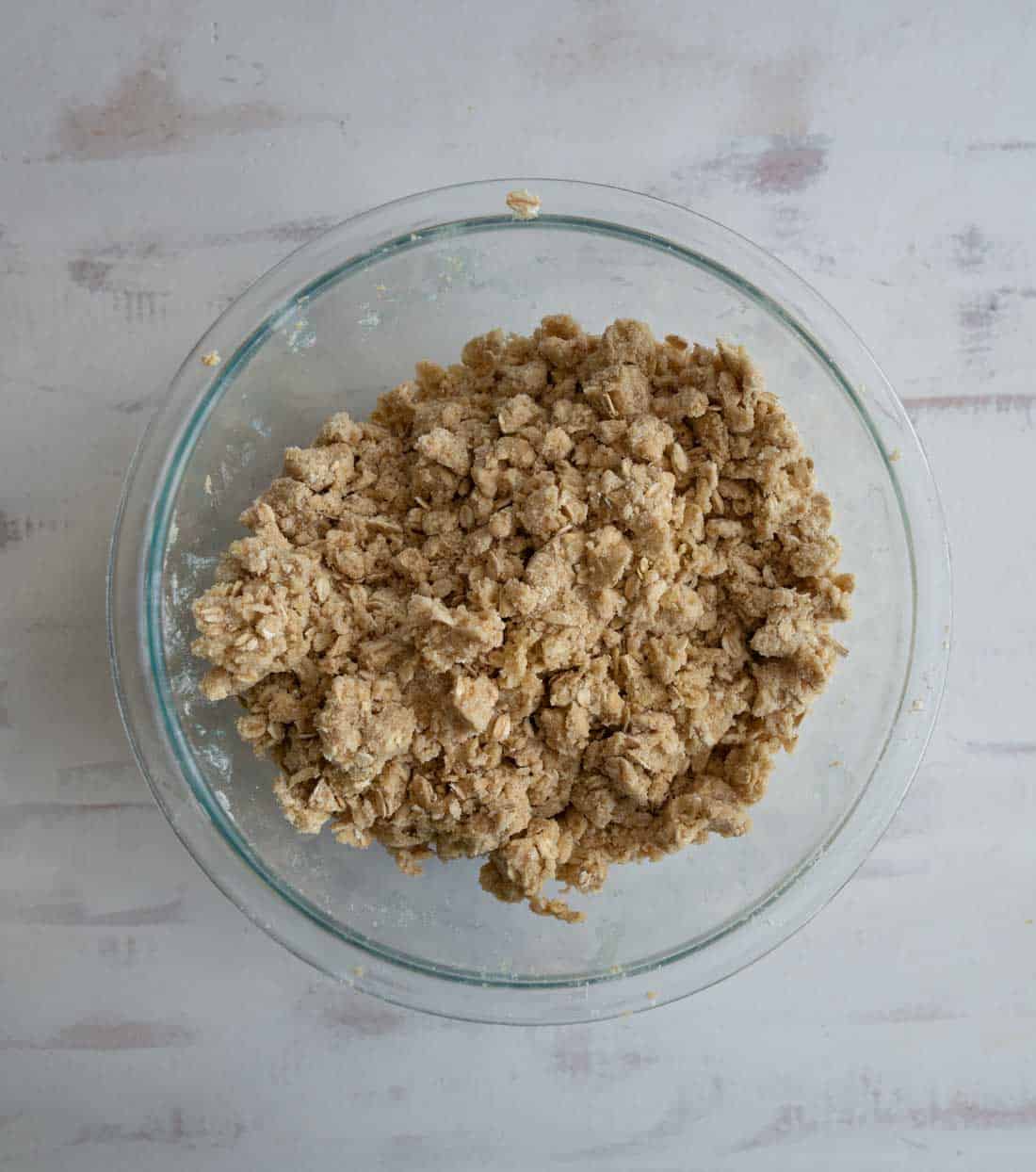 A glass bowl filled with coarse, crumbly dough sits on a light-colored, textured surface.
