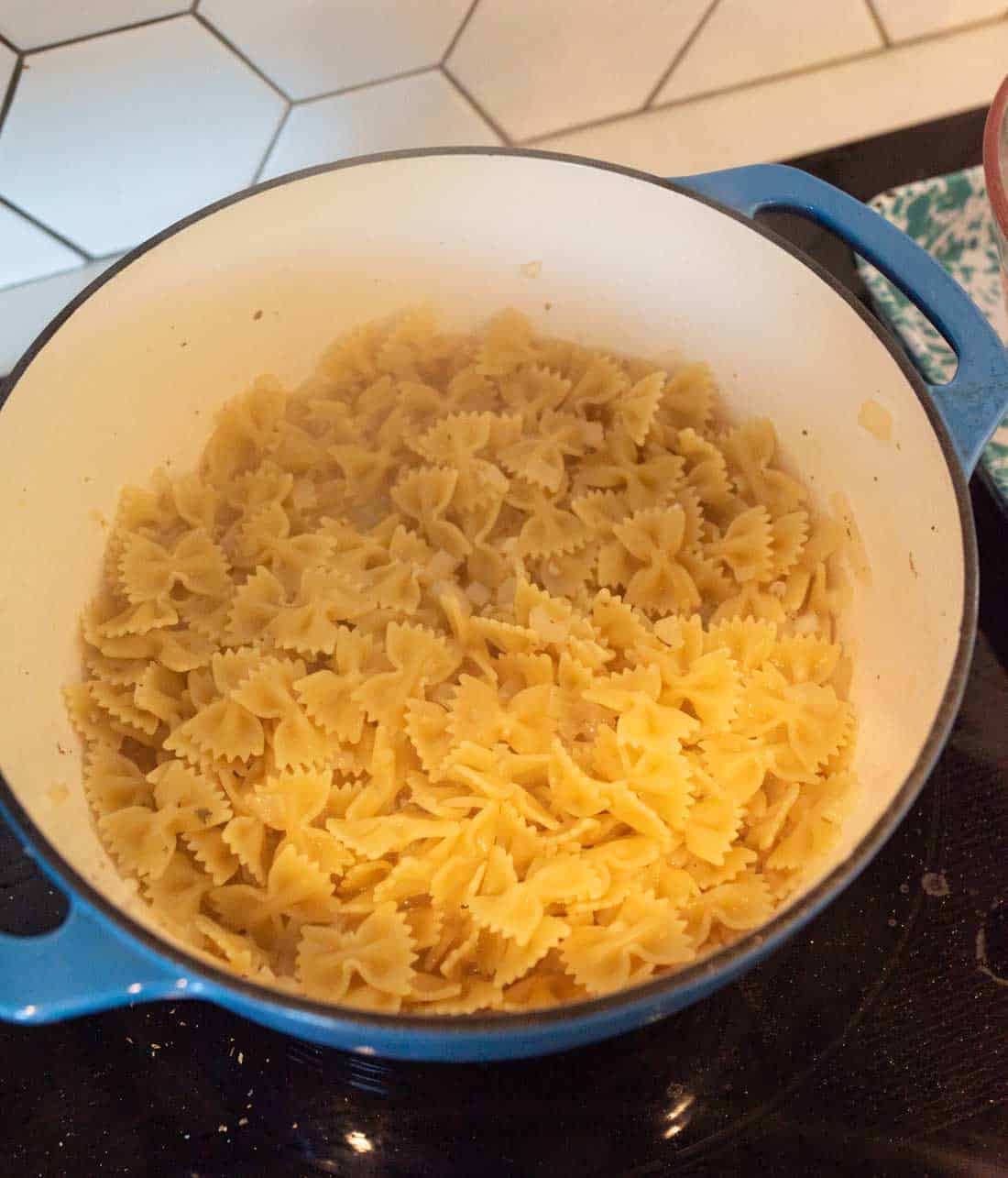 Cooked bowtie pasta in a blue pot on a stovetop, with a white tiled backsplash visible in the background. Steam rises from the pasta, indicating it is freshly cooked.