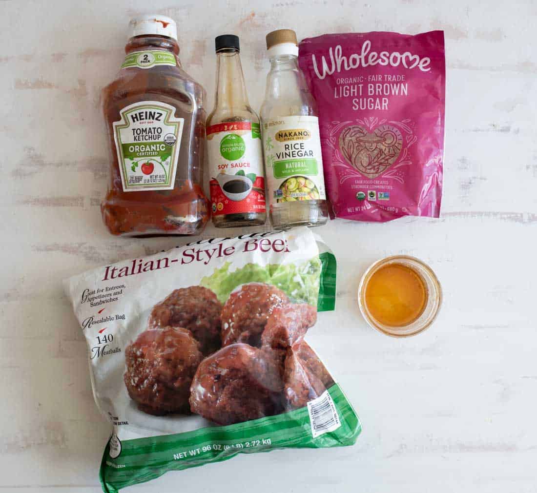 A bottle of ketchup, soy sauce, rice vinegar, a bag of light brown sugar, frozen Italian-style meatballs, and a small bowl of sauce are arranged on a white surface.