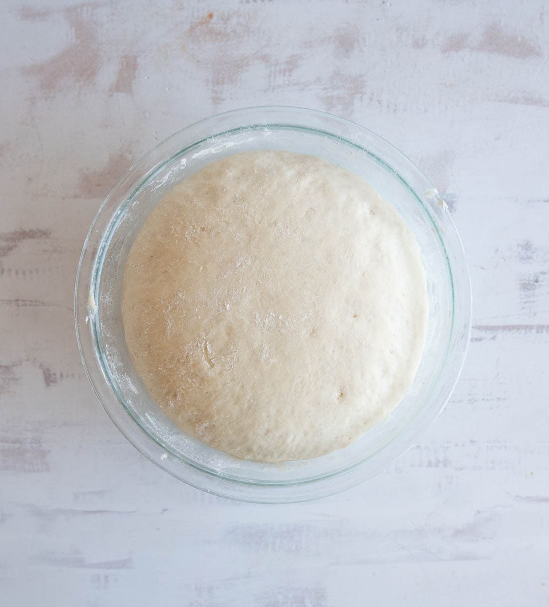 A glass bowl filled with risen bread dough sits on a light-colored, textured surface. The dough has expanded and appears soft and slightly puffy.