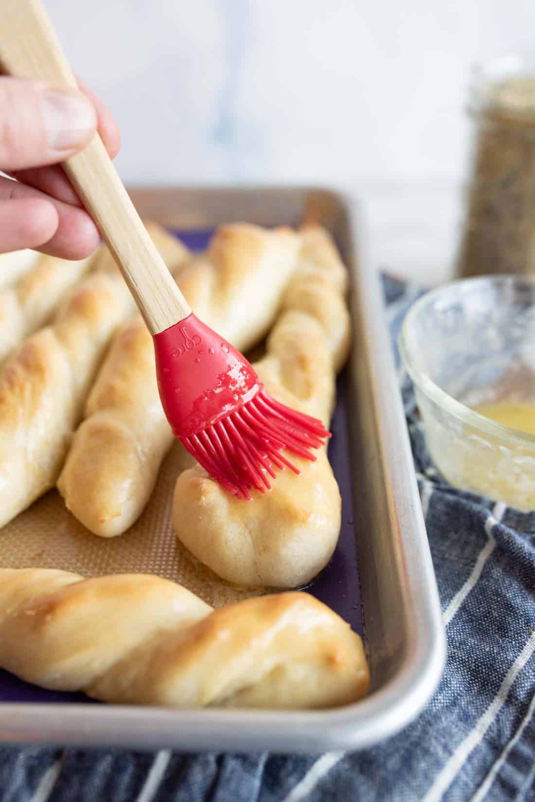 A hand uses a red silicone brush to apply butter to freshly baked breadsticks on a baking tray, with a small glass bowl of melted butter nearby.