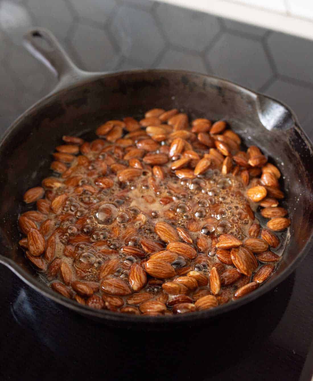 Whole almonds being roasted in a black cast iron skillet with bubbling oil or syrup, on a stovetop with a dark hexagonal tile backsplash in the background.