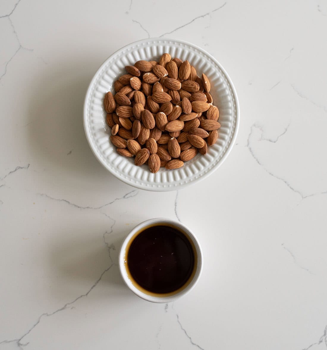 A white bowl filled with raw almonds and a small white cup of dark liquid, possibly vanilla extract, are placed on a white marble surface.