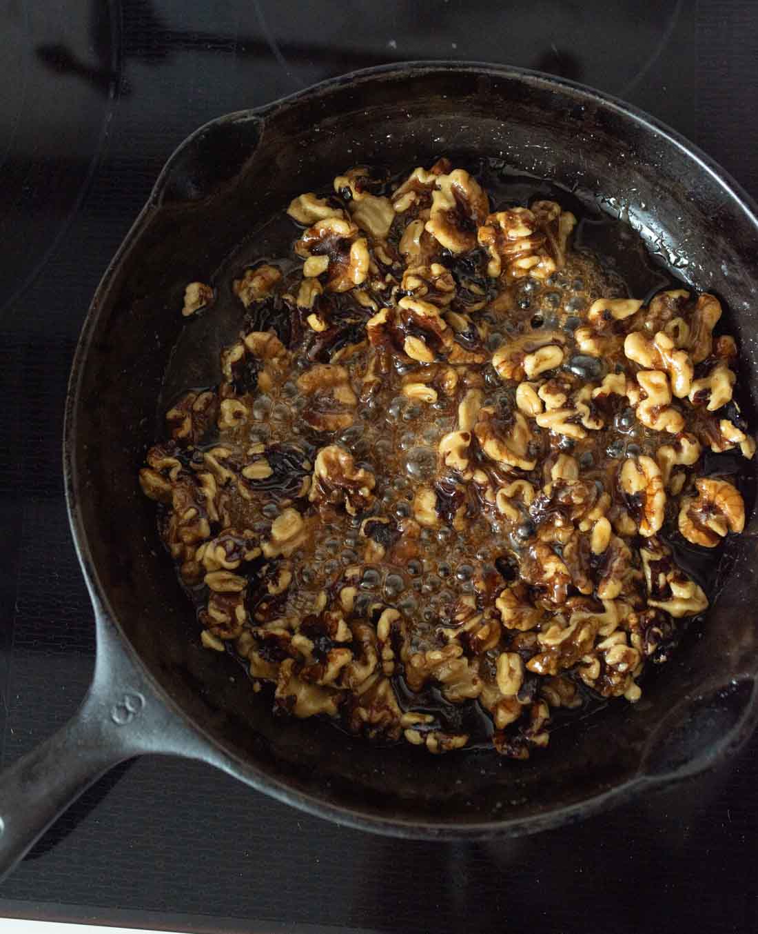 A cast iron skillet on a stovetop containing walnuts being caramelized in bubbling sugar or syrup.