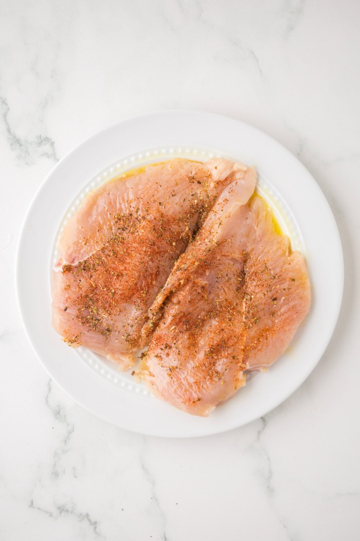 Raw, seasoned chicken breasts are arranged on a white plate. The chicken is coated with a blackened chicken spice blend and herbs, sitting on a white marble surface.